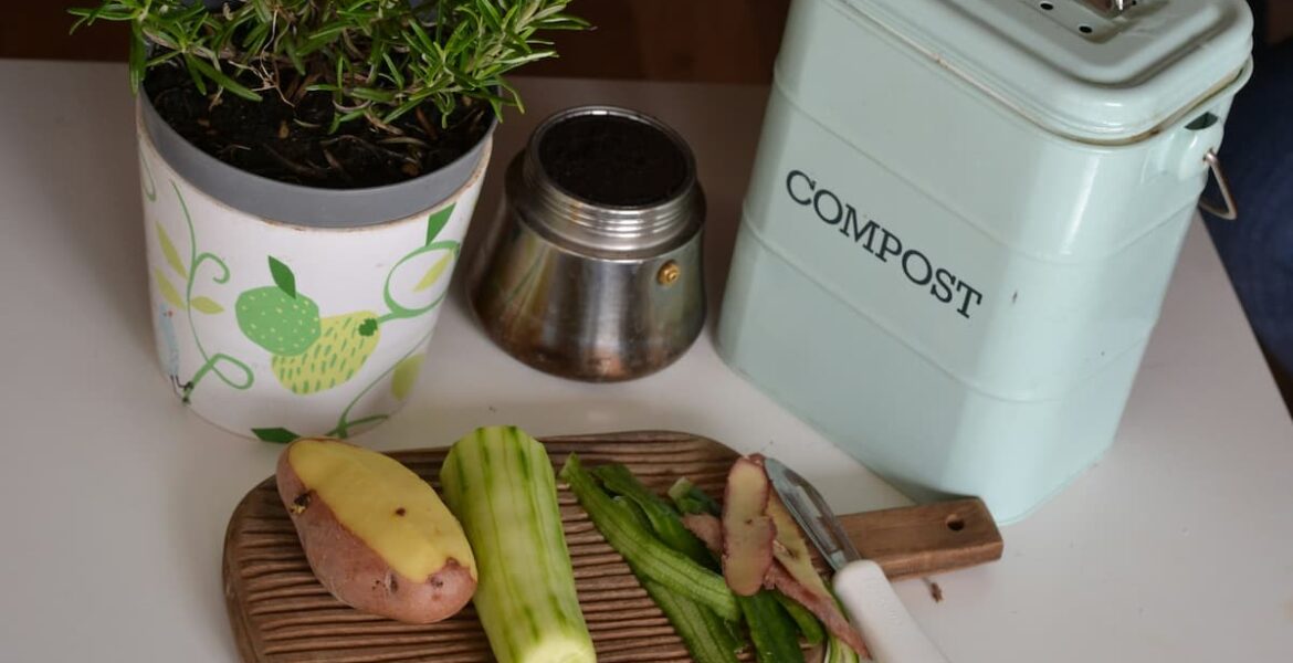 Recortes de vegetales sobre una tabla junto a un contenedor de compost de cocina y una maceta con planta aromática.