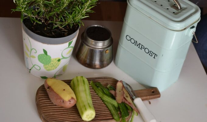 Recortes de vegetales sobre una tabla junto a un contenedor de compost de cocina y una maceta con planta aromática.