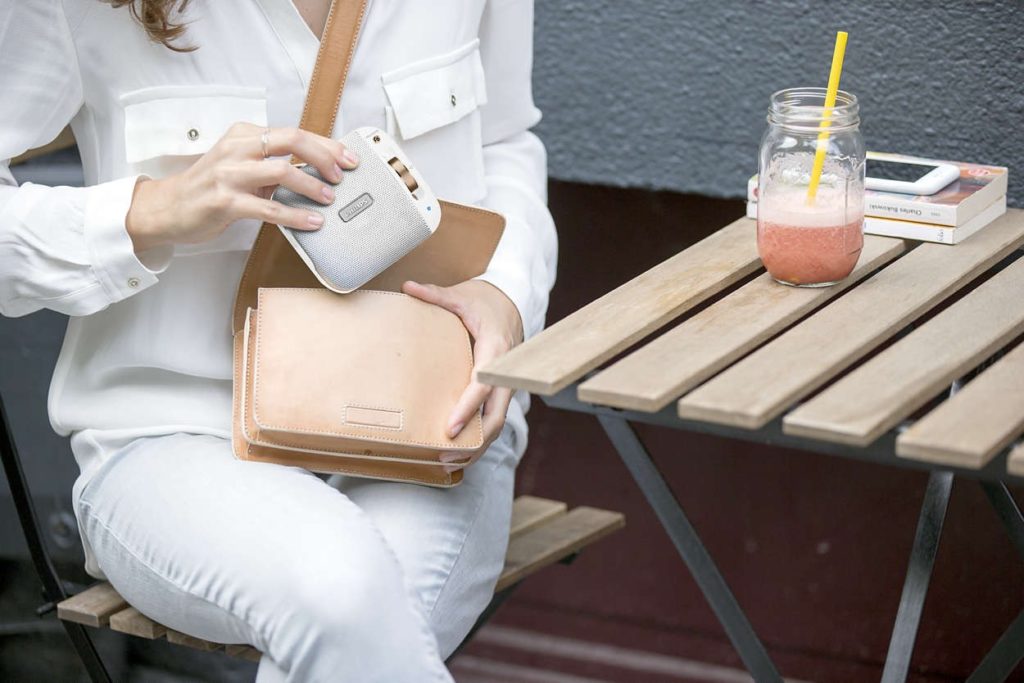 Mujer guarda un parlante portátil compacto en su bolso mientras toma un batido en una terraza.