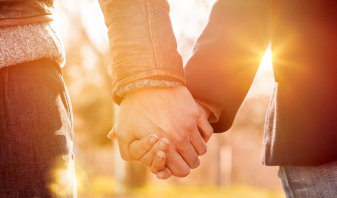 Pareja tomada de la mano al atardecer, con luz cálida y fondo natural desenfocado.
