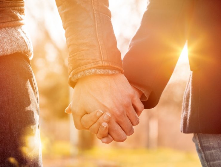 Pareja tomada de la mano al atardecer, con luz cálida y fondo natural desenfocado.
