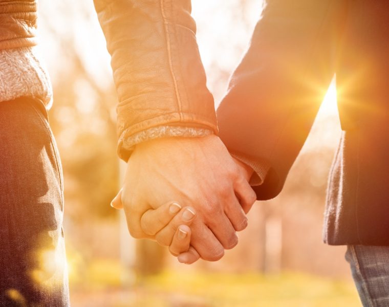 Pareja tomada de la mano al atardecer, con luz cálida y fondo natural desenfocado.