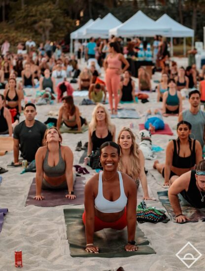 Personas practicando yoga sobre la arena en un evento Daybreaker frente al mar.