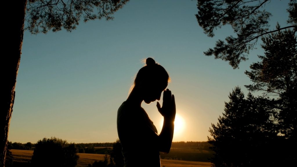 Mujer en silueta meditando al atardecer con las manos en posición de oración.
