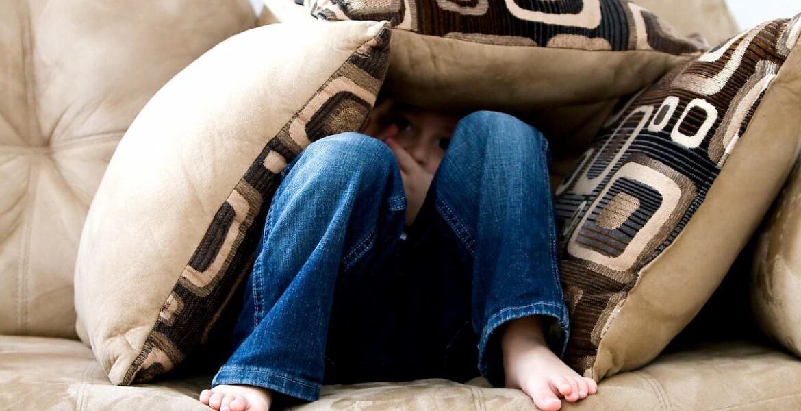Niño escondido entre cojines en un sillón por miedo.