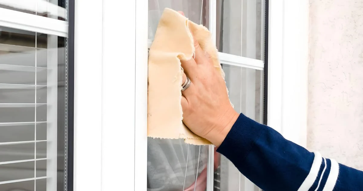 Mano con un paño amarillo limpiando el cristal de una ventana de marco blanco