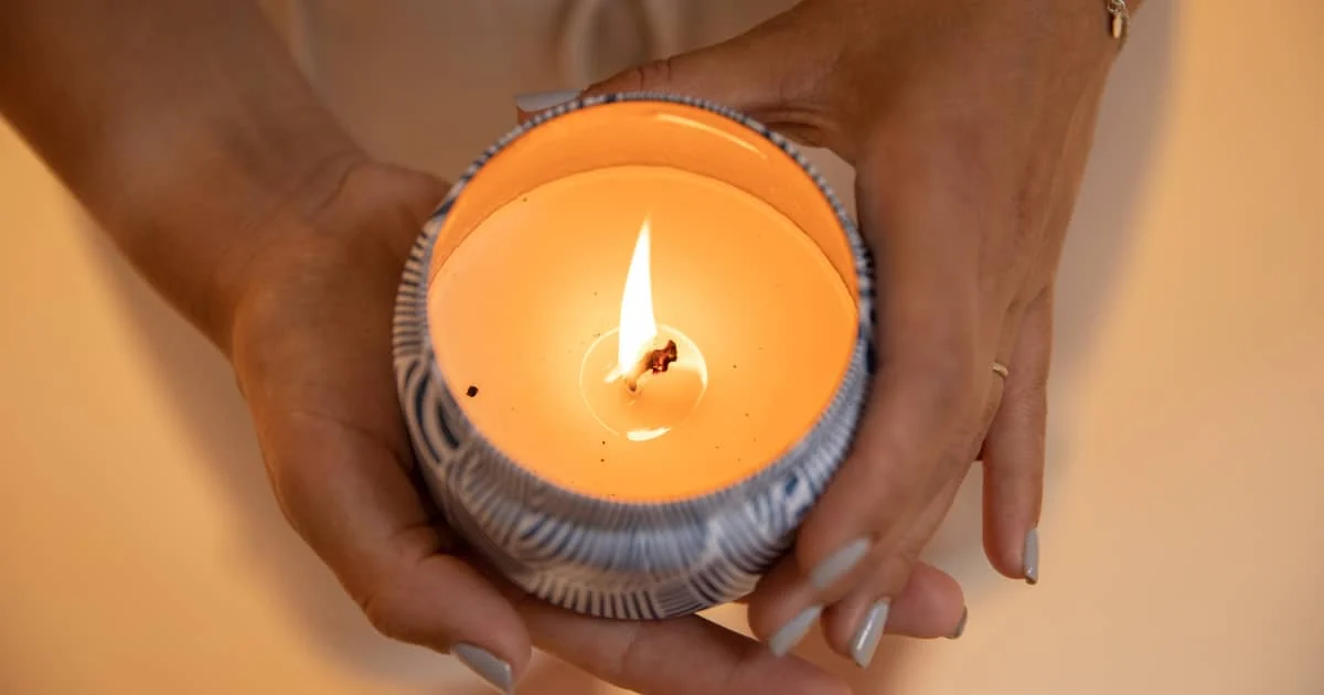 Manos con las uñas pintadas sostienen una vela encendida en un recipiente azul estampado