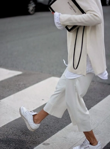Look de street style de una mujer combinando un pantalón blanco culotte con tenis blancos y un suéter de punto, llevando una bolsa de la marca Balenciaga.