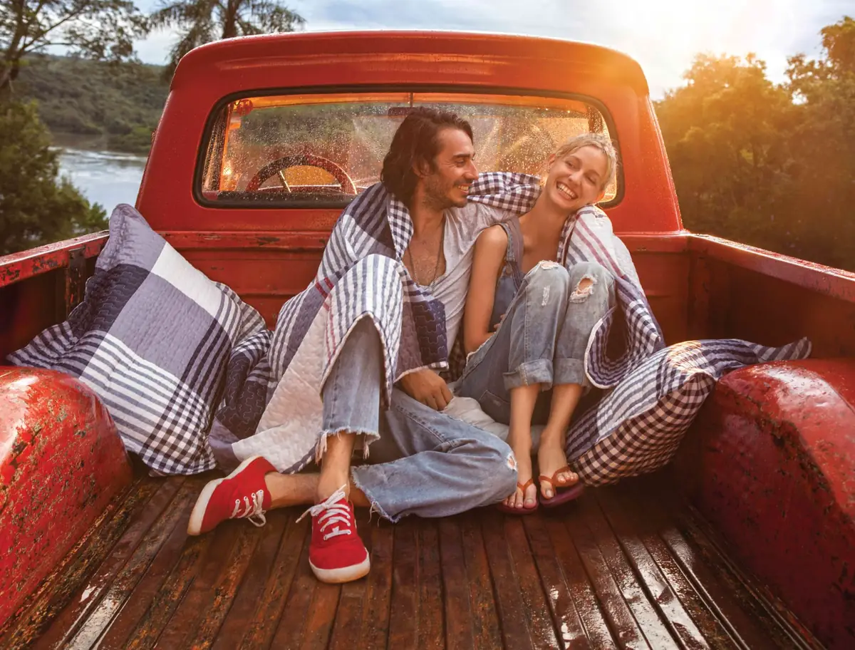 Pareja joven sonriendo y abrazada en la parte trasera de una camioneta roja, envuelta en una manta, al atardecer en un entorno natural.