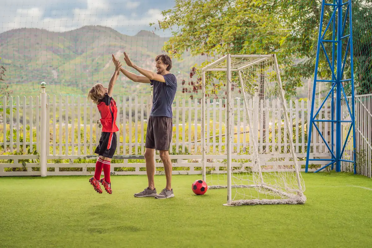 Padre e hijo chocando las manos (high-five) en un campo de fútbol, representando una experiencia memorable como regalo para el Día del Padre.