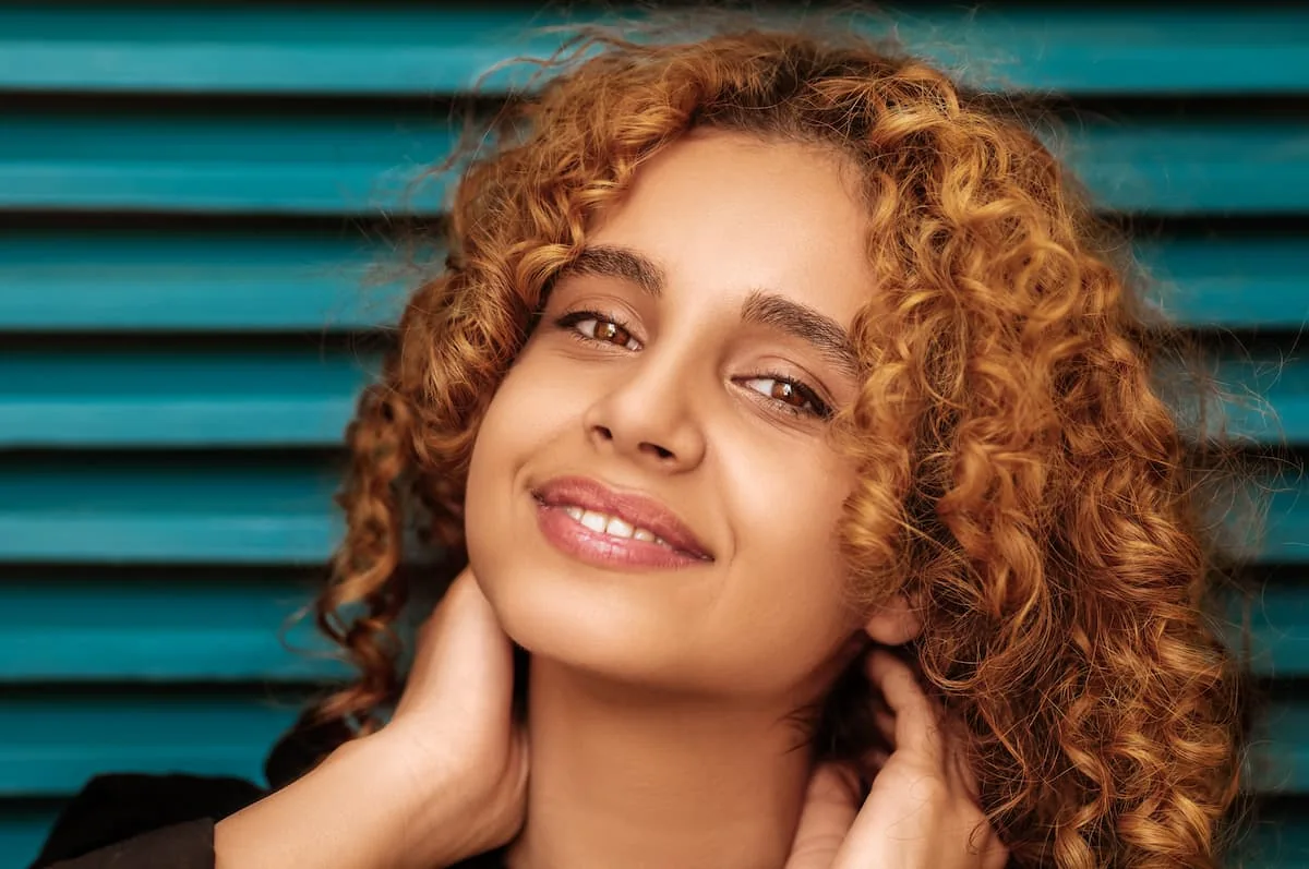 Cabello rizado definido, teñido con un balayage en tonos castaño cobrizo y caramelo que iluminan sus facciones.