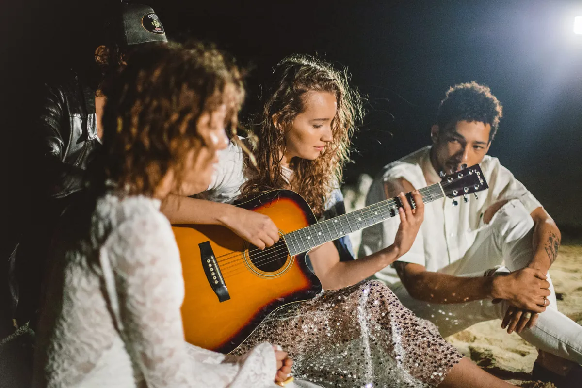 Aprender a tocar guitarra acústica desde cero para principiantes Grupo de amigos en una reunión nocturna al aire libre, con una joven mujer tocando la guitarra acústica y cantando.