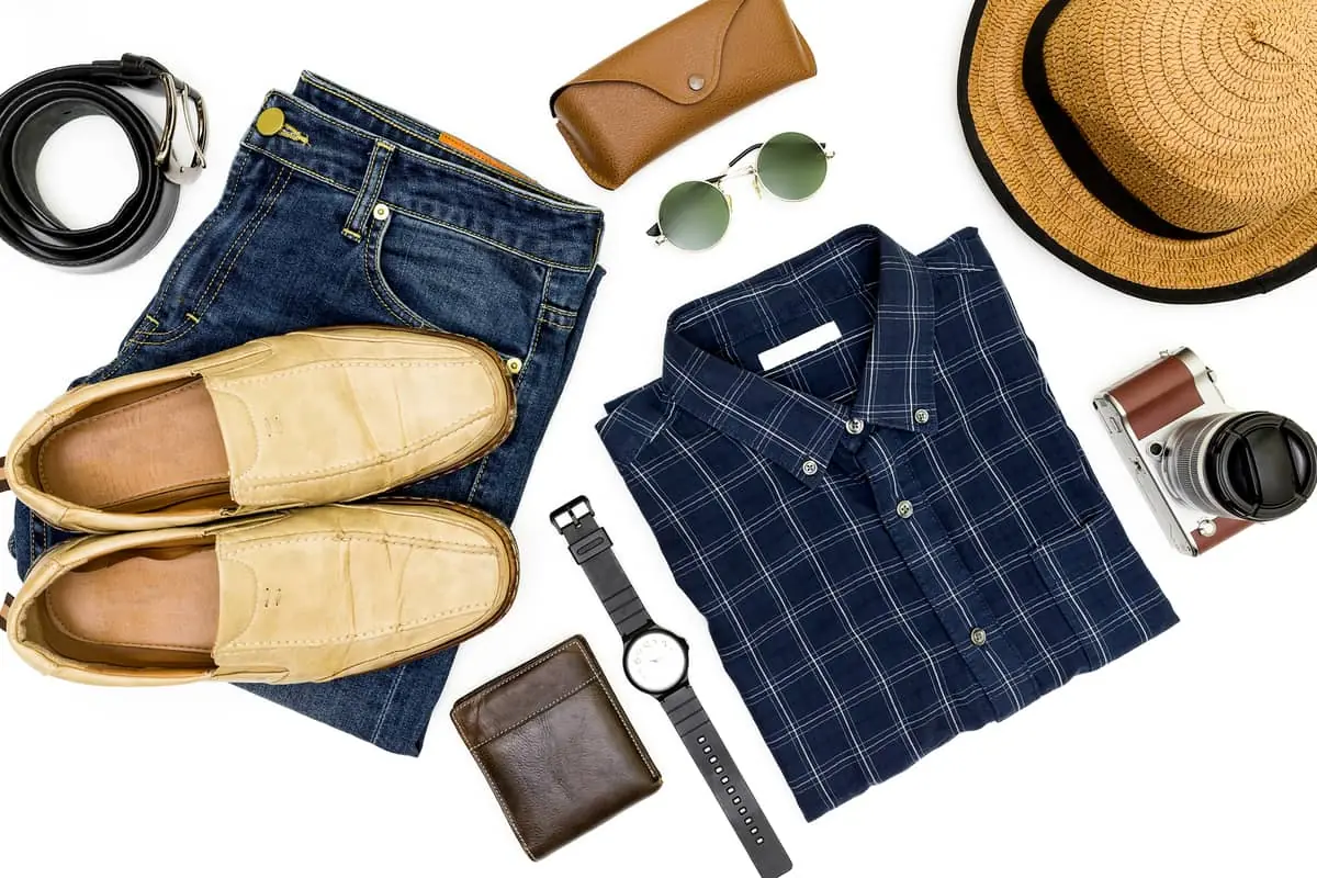 Flat lay de un outfit casual para hombre, incluyendo jeans de mezclilla, una camisa de cuadros azul, mocasines de cuero beige y accesorios de viaje como sombrero, gafas de sol y cámara.