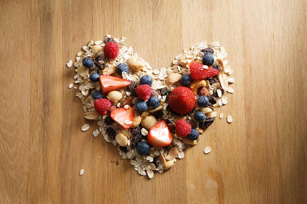Corazón formado con granola, avena, fresas, moras y nueces sobre fondo de madera clara.