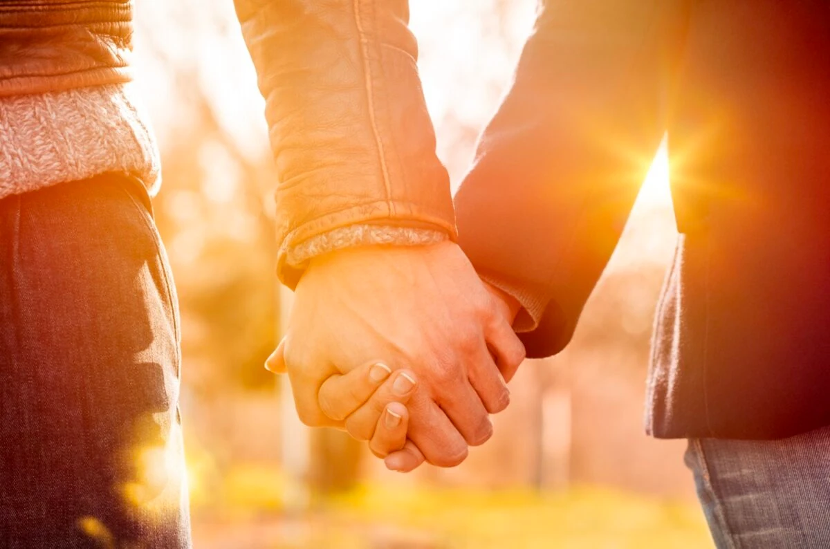 Pareja tomada de la mano al atardecer, con luz cálida y fondo natural desenfocado.