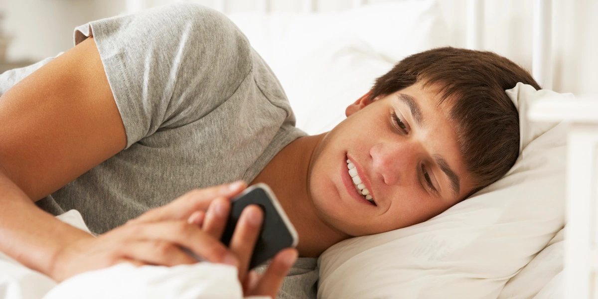 Joven acostado en la cama mirando el celular, revisando redes sociales.