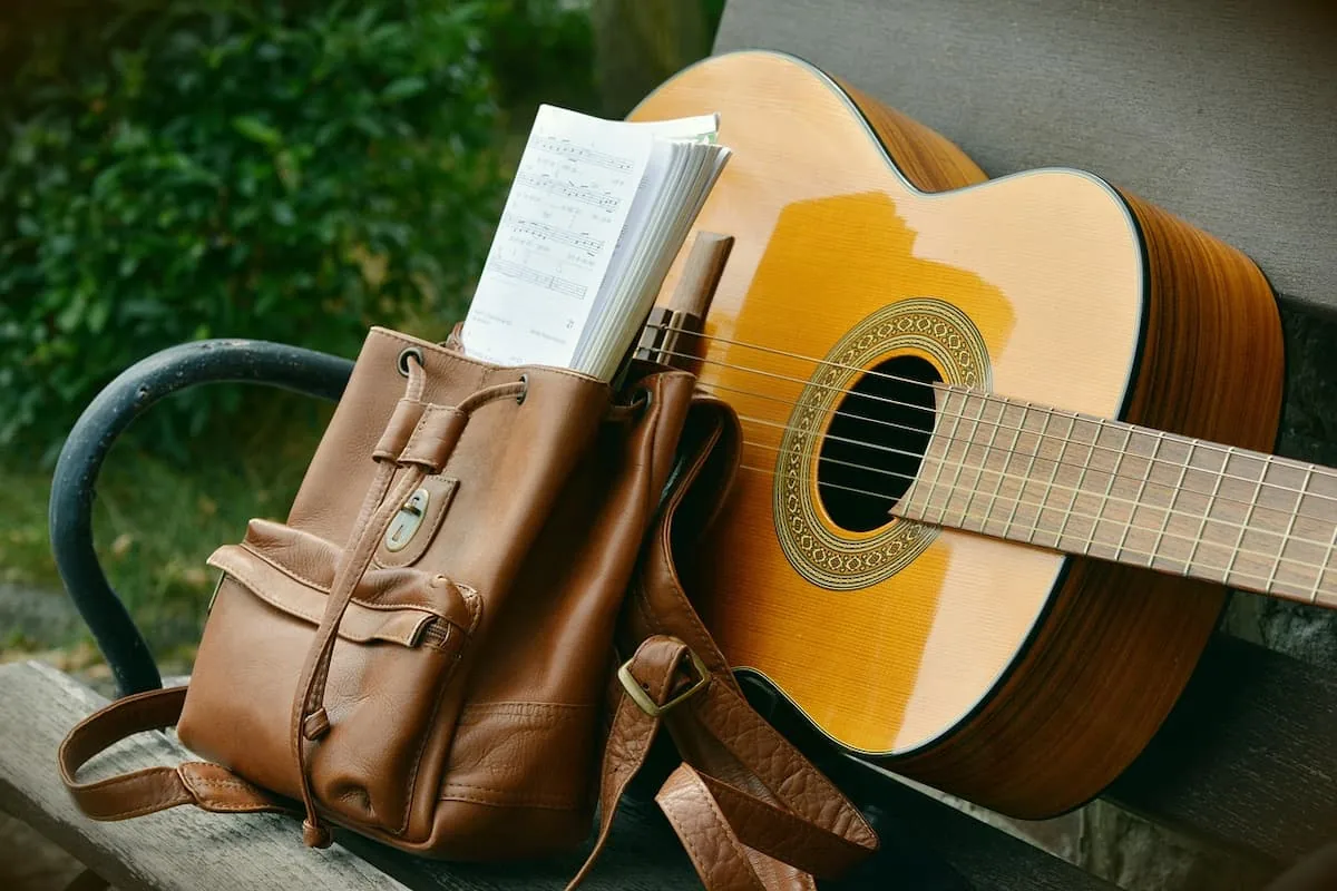 El Primer Paso para Aprender a Tocar Guitarra Guitarra acústica apoyada en un banco junto a una mochila con partituras, representando los elementos esenciales para aprender a tocar desde cero.