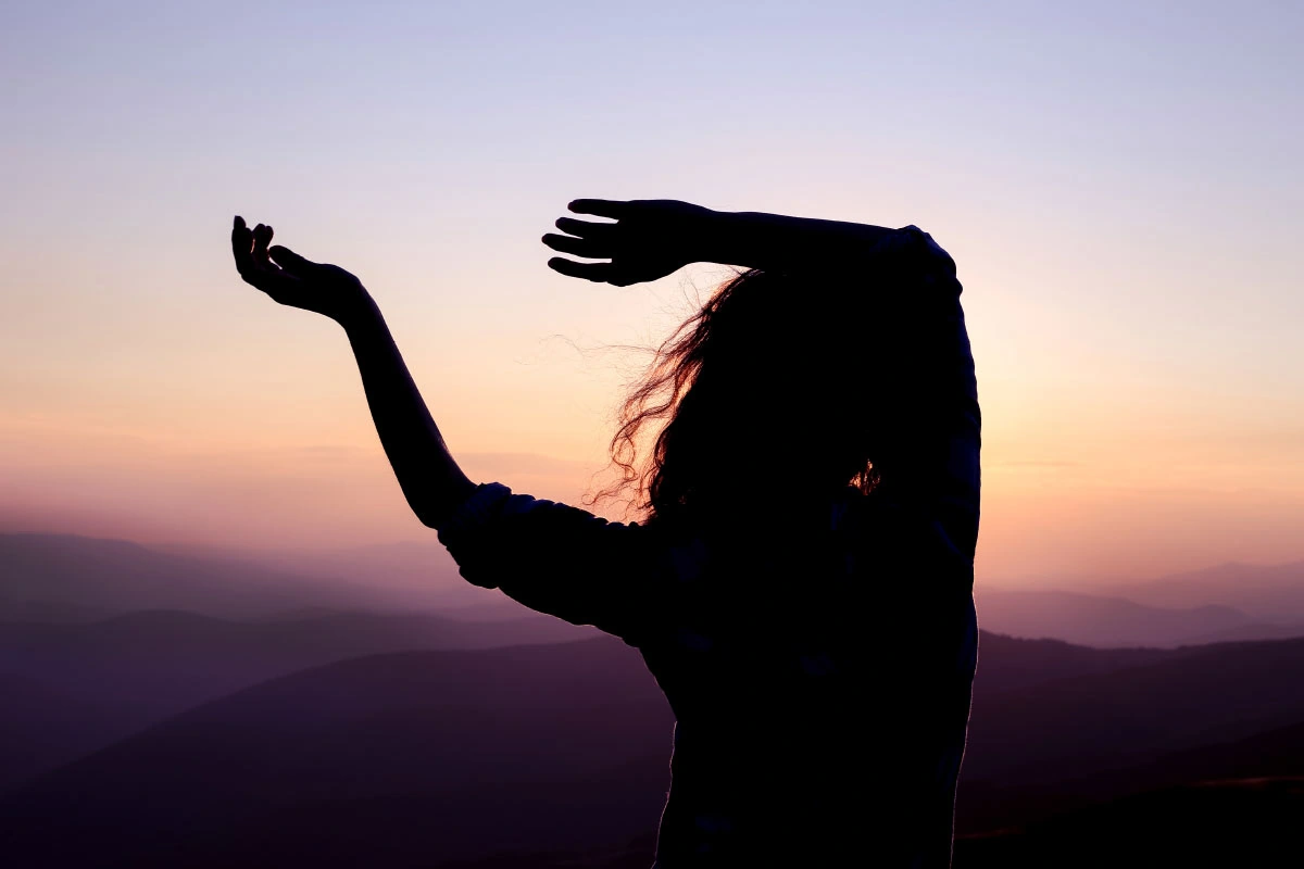 Meditación en movimiento: la danza libre que armoniza cuerpo y espíritu Silueta de una mujer bailando al atardecer, con el cielo violeta y montañas al fondo, simbolizando libertad y conexión interior.
