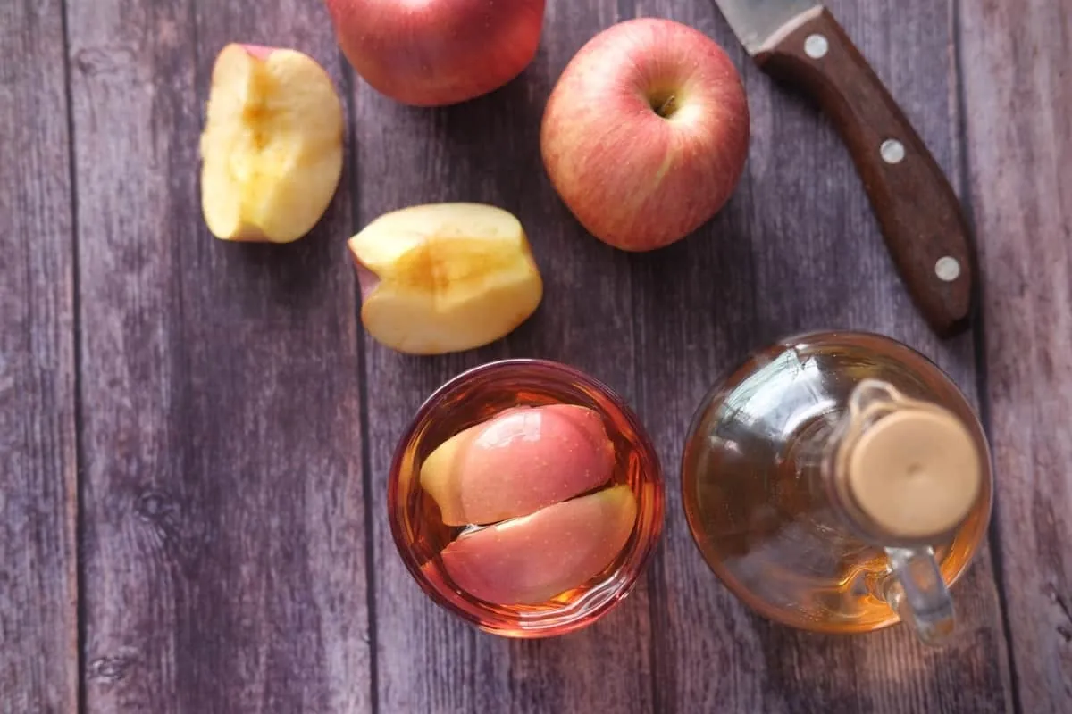 Mezclar vinagre de manzana con agua y aplicarlo puede ayudar a equilibrar el pH del cuero cabelludo. Trozos de manzanas rojas con un recipiente de vinagre de manzana