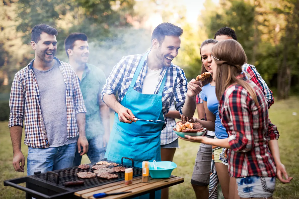 La parrilla: el pretexto perfecto para reunirte con amigos. Grupo de amigos jóvenes riendo y disfrutando de una parrillada o carne asada al aire libre alrededor de un asador.
