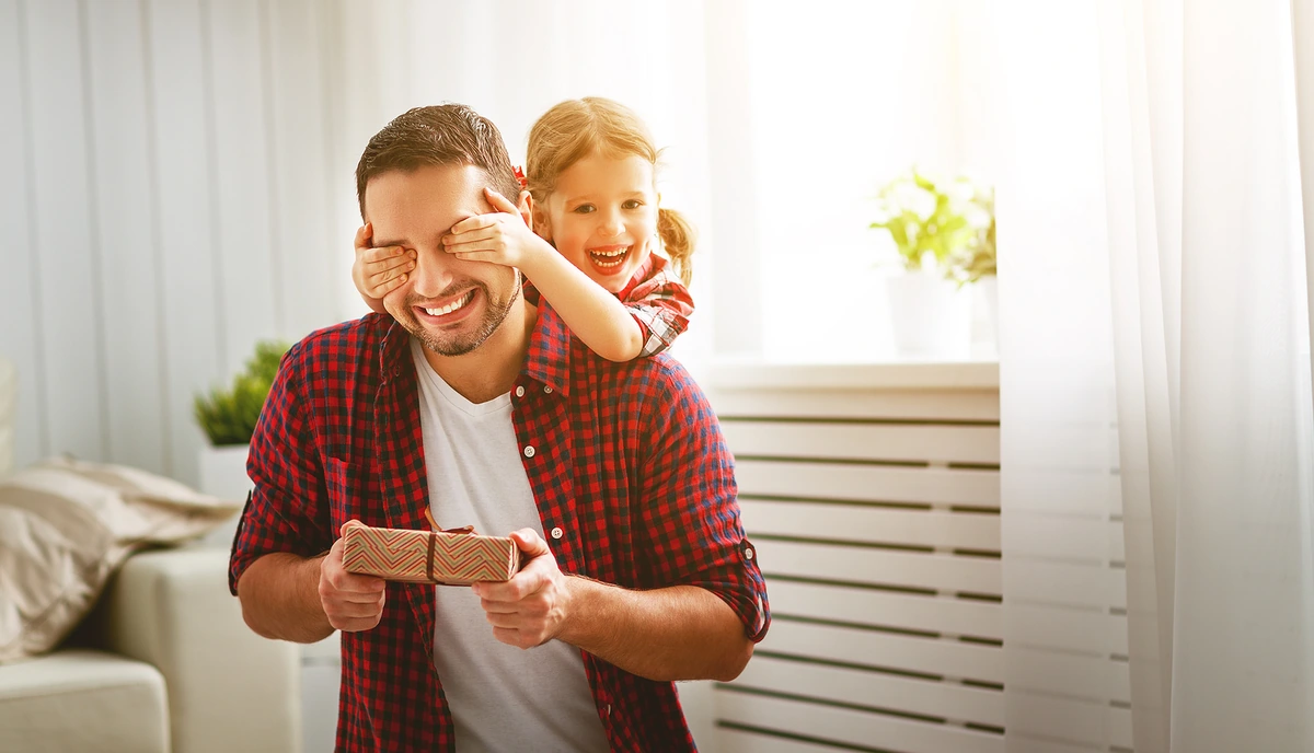 Niña pequeña cubriendo los ojos de su papá para sorprenderlo con un regalo, ilustrando ideas de obsequios originales para hombres y padres.