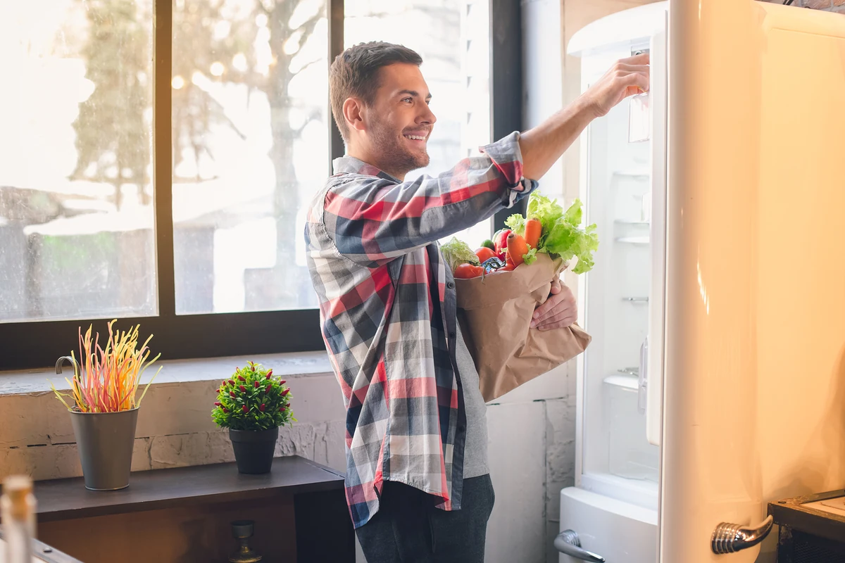 Hombre sonriente guardando verduras frescas en el refrigerador de su cocina en Estados Unidos.