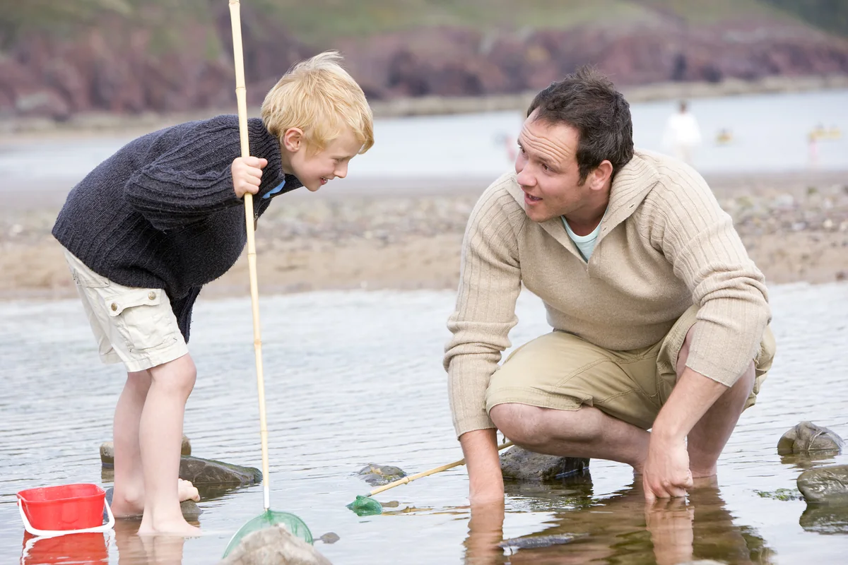 Padre e hijo explorando juntos una poza en la playa con una red, representando el regalo de una aventura para el Día del Padre.