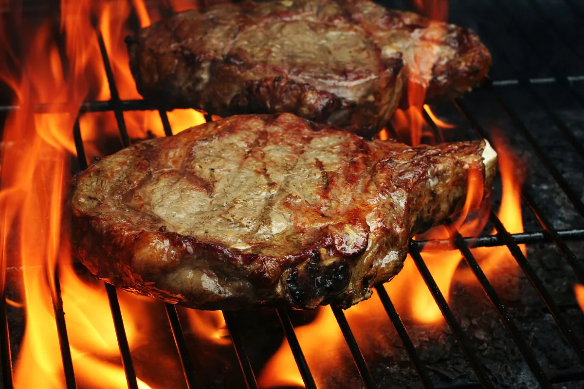 Cómo Asar el Corte de Carne Perfecto a la Parrilla Dos jugosos cortes de carne ribeye cociéndose a fuego directo en una parrilla.