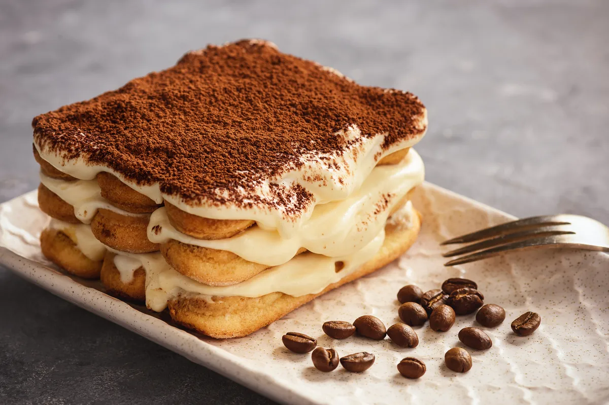 Postre de vainilla con crema pastelera y cacao en polvo, servido en plato rectangular con granos de café y un tenedor.