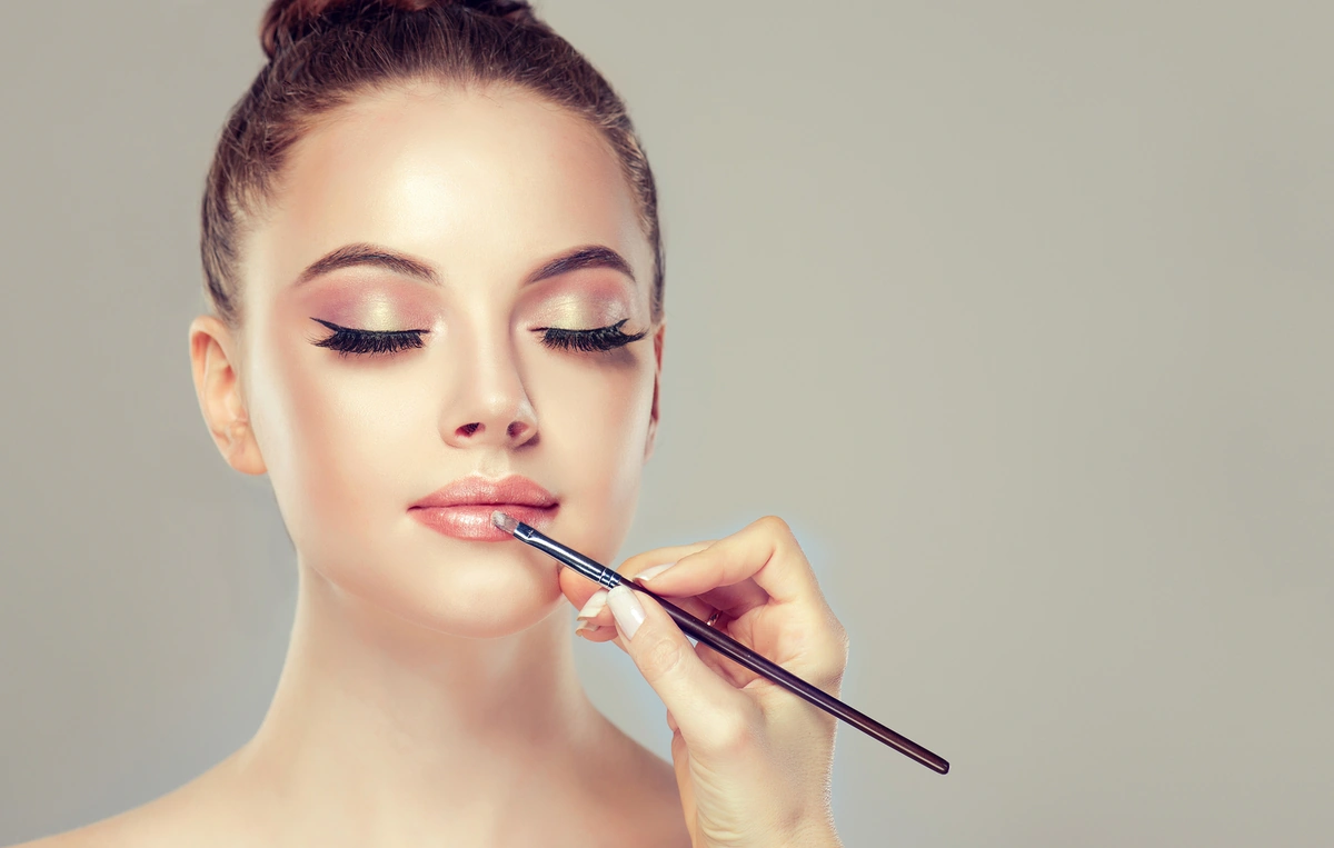 Aprender a delinear los labios permite que los labiales se aprovechen mejor. Mujer joven delineando su labios con un pincel luego de terminar su rutina de maquillaje