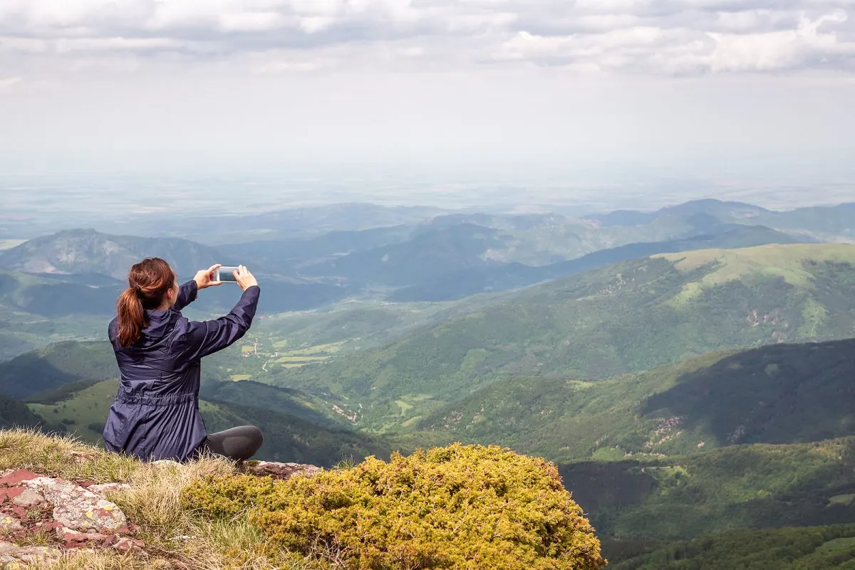 Mujer tomando una foto del paisaje montañoso con su celular desde la cima de una montaña.