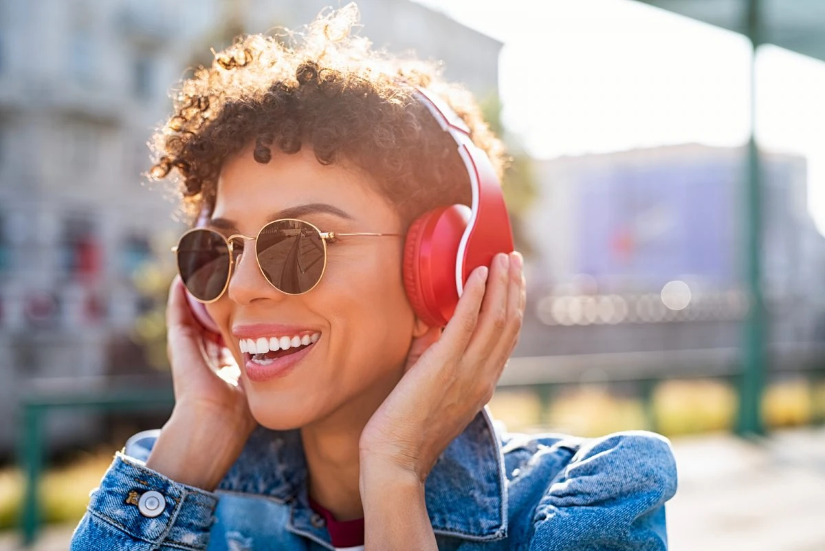 Mujer joven escuchando música con auriculares inalámbricos rojos al aire libre, sonriendo bajo el sol.