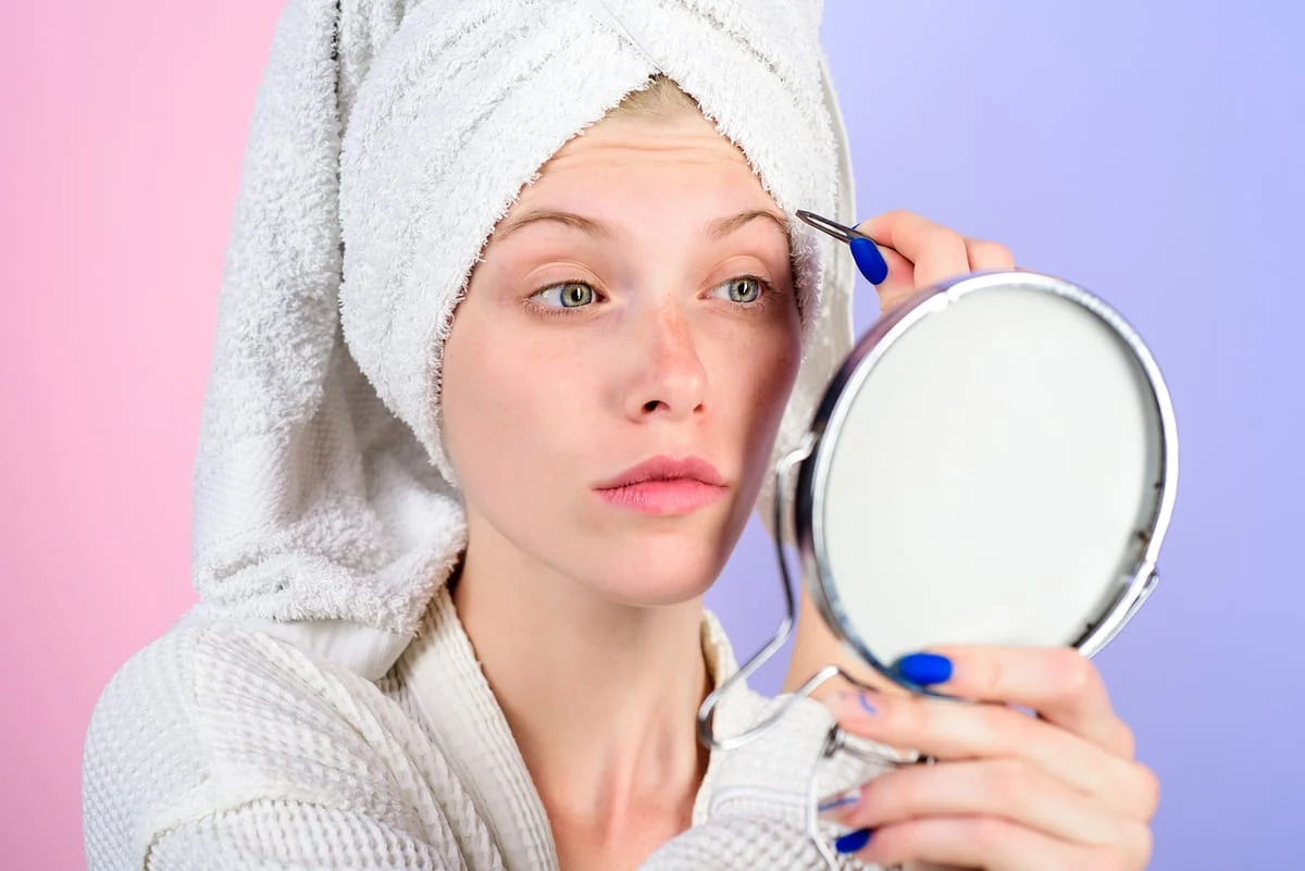Mujer en bata de baño perfilando sus cejas con pinzas frente a un espejo para enmarcar la mirada.
