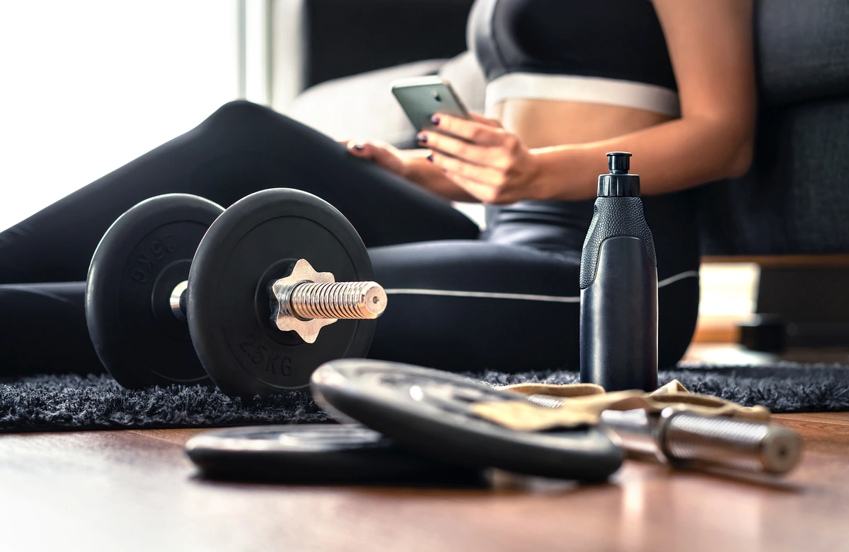 Entrenamiento en casa con rutinas de YouTube Mujer con ropa deportiva revisando el celular junto a pesas y botella de agua, ejemplo de entrenamiento en casa con YouTube