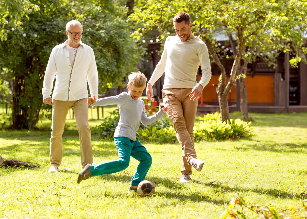 Tres generaciones de una familia, abuelo, padre e hijo, jugando fútbol juntos en el jardín, una imagen que celebra el legado familiar en el Día del Padre.