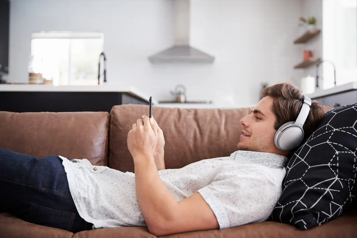 Hombre joven recostado en un sofá escuchando música con auriculares inalámbricos mientras usa su teléfono.