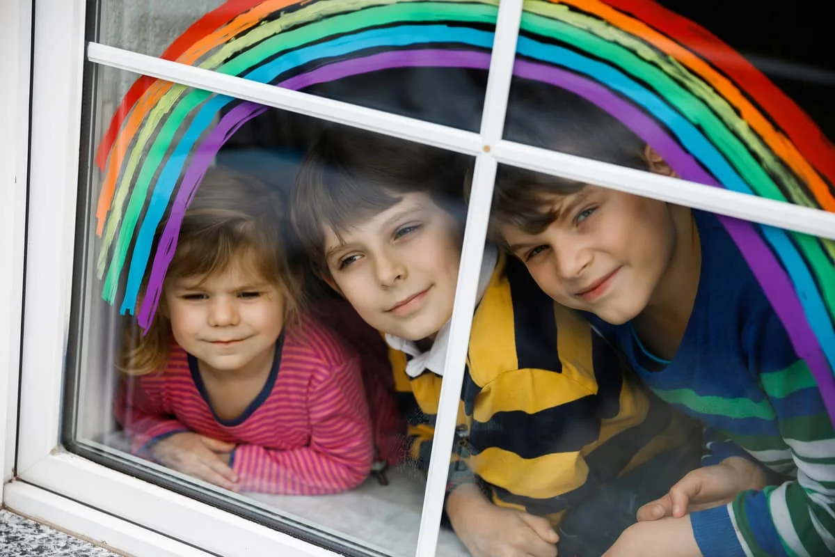 Tres niños mirando por la ventana con un arcoíris pintado, símbolo de esperanza.