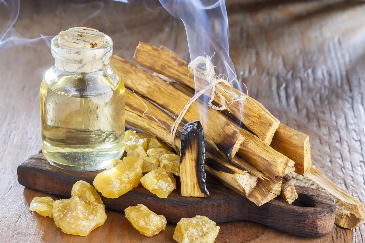 Palo santo encendido con humo, acompañado de resinas y frasco de aceite esencial sobre tabla de madera.
