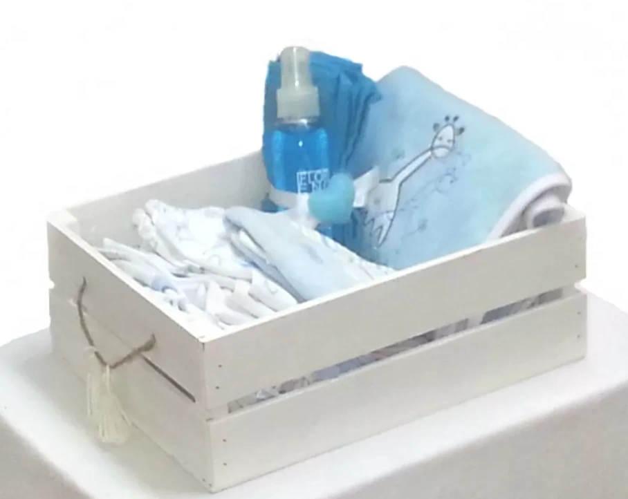 Canasta de regalo para bebé en una caja de madera blanca, con ropa, una manta de jirafa y productos de aseo.