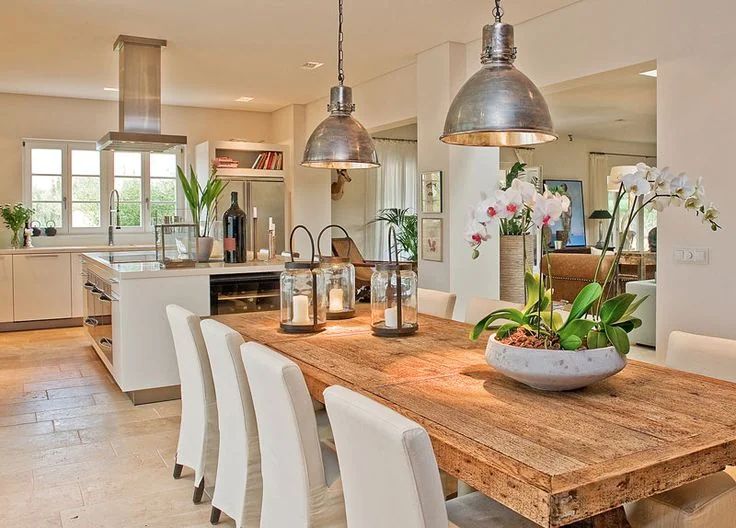 Cocina comedor de concepto abierto con una gran mesa de madera rústica, sillas blancas y lámparas industriales de metal, creando un estilo rústico elegante.