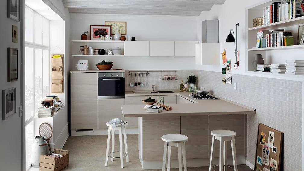 Cocina pequeña moderna en tonos claros con barra en L y taburetes blancos, organizada con estantes abiertos y buena iluminación natural.