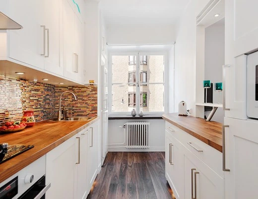 Cocina pequeña alargada con muebles blancos, encimeras de madera y salpicadero de mosaicos de colores.