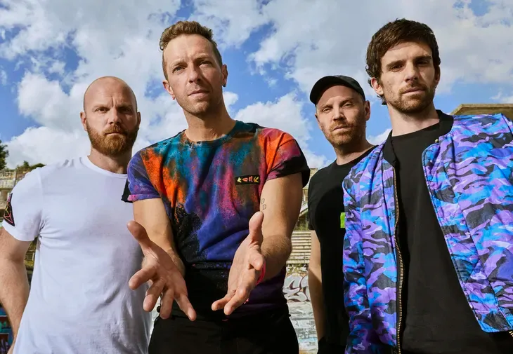 Los cuatro miembros de Coldplay posando juntos al aire libre con cielo azul

