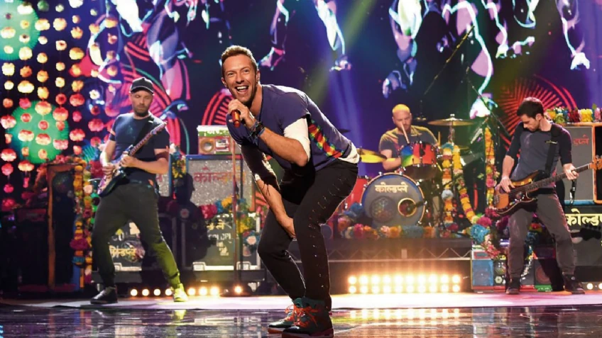 Escena vibrante del concierto de Coldplay donde Chris Martin interactúa con el público, acompañado por Jonny Buckland (guitarra), Guy Berryman (bajo) y Will Champion (batería).