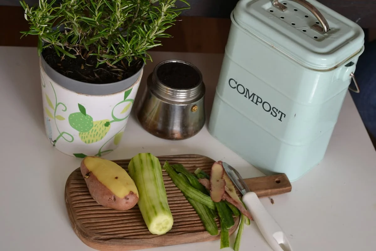 Recortes de vegetales sobre una tabla junto a un contenedor de compost de cocina y una maceta con planta aromática.