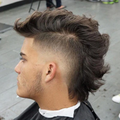 Mullet con Fade: La Fusión Perfecta de Estilos Modernos Hombre joven con un corte de pelo mullet moderno, caracterizado por un degradado (fade) muy marcado en los laterales y la nuca, contrastando con el cabello largo y texturizado en la parte superior y trasera.