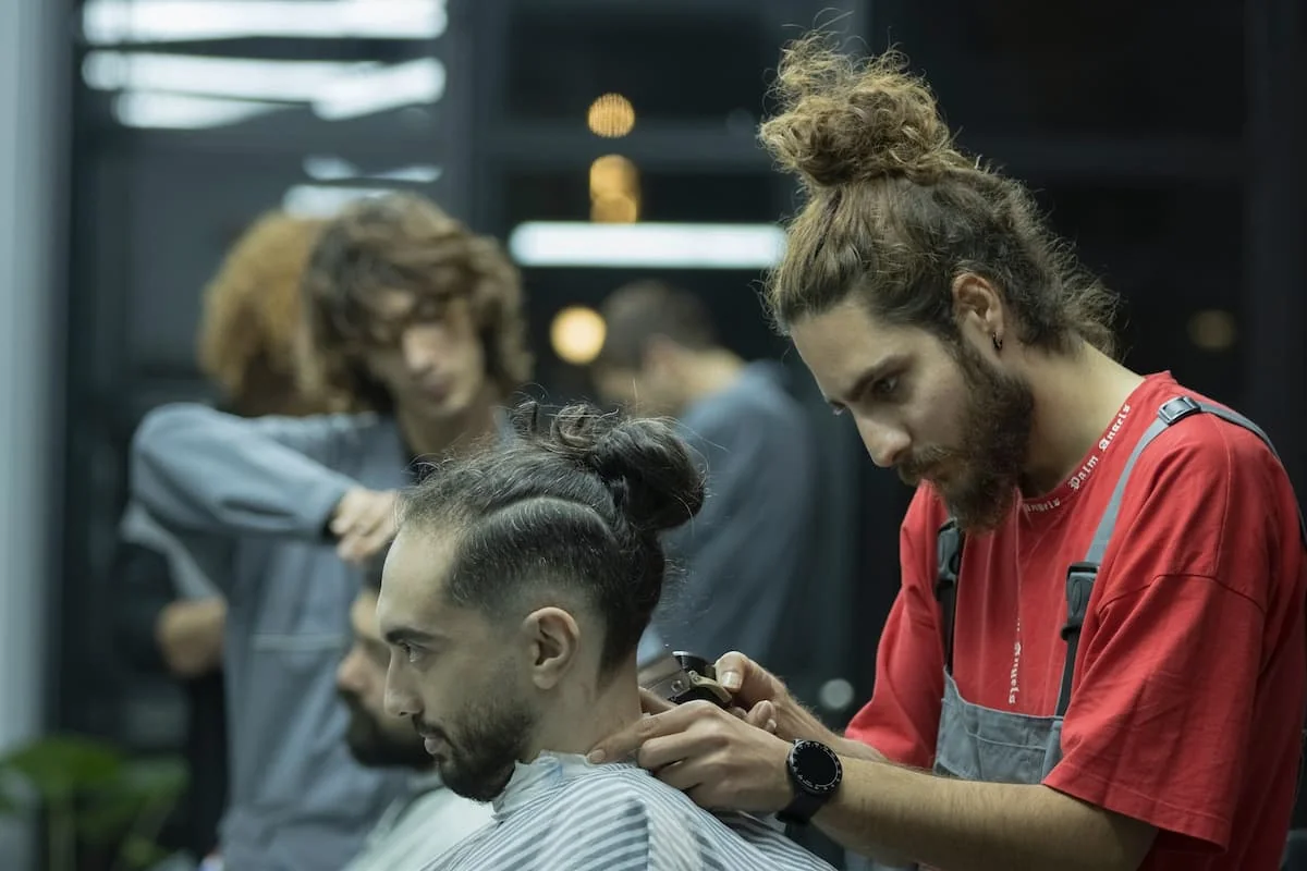 Barbero profesional concentrado, usando máquina para hacer un corte de pelo degradado o 'fade' a un cliente con un moño alto, representando la precisión en la barbería moderna.