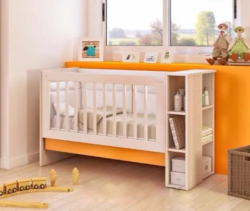Cuna de bebé blanca y madera con estantería de almacenaje integrada en el extremo, ideal para cuartos pequeños y con pared naranja de fondo.
