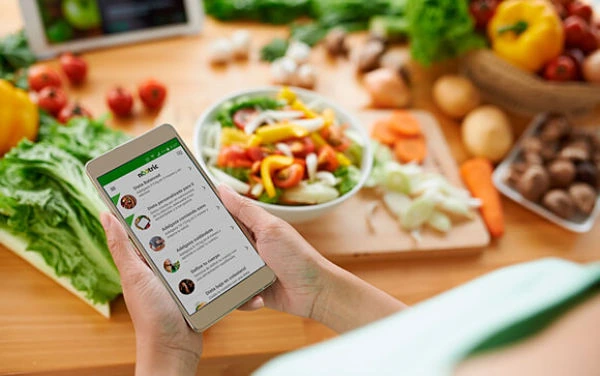 Planificación de comidas con IA Descripción: Persona usando una aplicación de IA en su celular para la planificación de comidas y la lista de compras, simplificando la organización del hogar con ingredientes frescos de fondo.