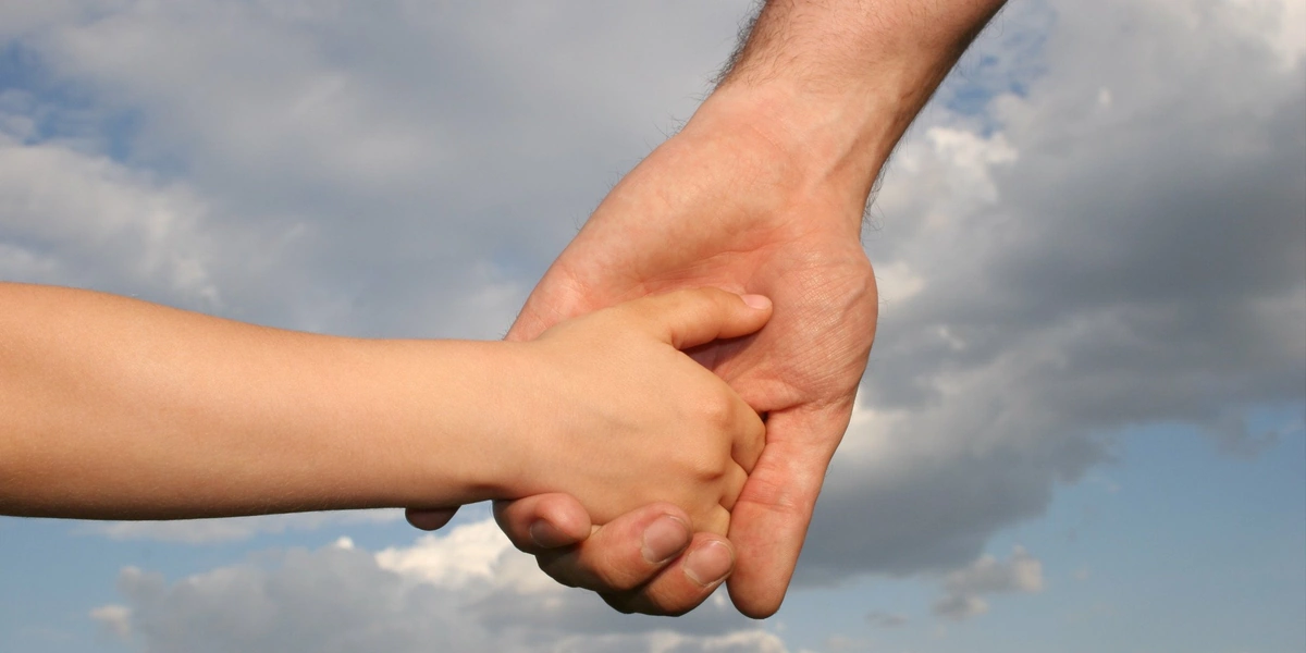 Mano de un adulto sujeta la de un niño sobre un cielo con nubes